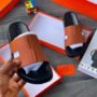 Brown leather slide sandals with black sole, presented in an open orange box with a hand holding one sandal, exuding luxury and comfort.