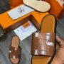 Brown croc-effect slide sandals with black sole, presented in an orange box with a hand holding one sandal.