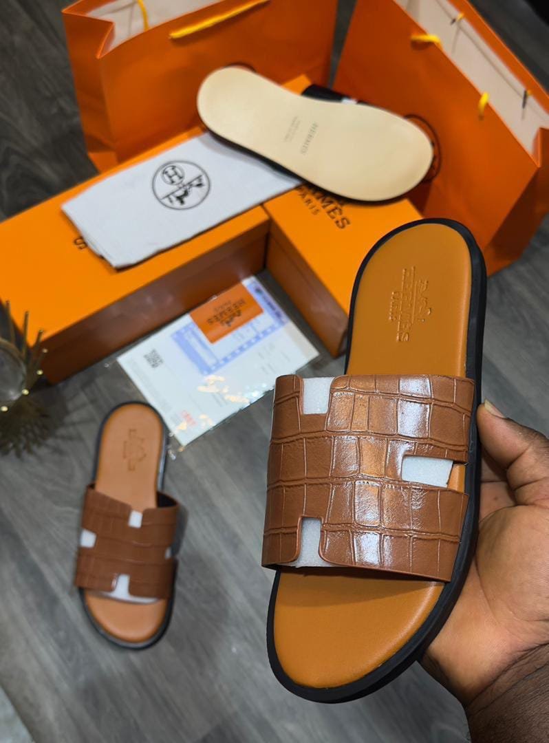Brown croc-effect slide sandals with black sole, presented in an orange box with a hand holding one sandal.