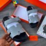 A pair of luxury grey slides with black sole, presented in an open orange box with a hand holding one slide, showcasing the premium quality and comfort of Splendourman shoes.
