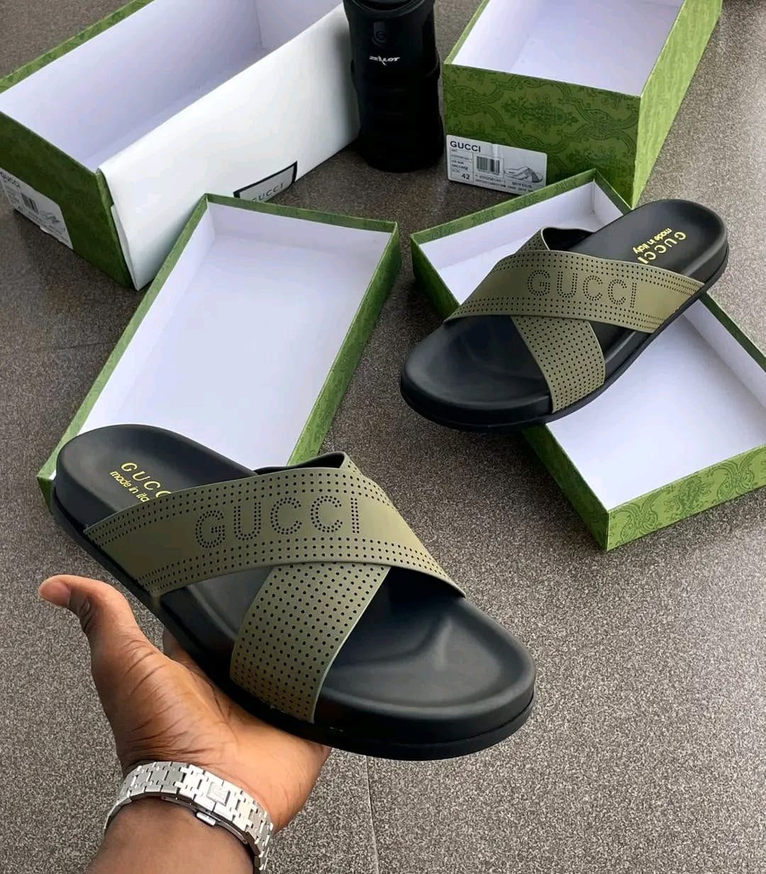 A hand holding an olive green slide sandal with a black sole and Gucci-inspired design, with shoe boxes in the background.