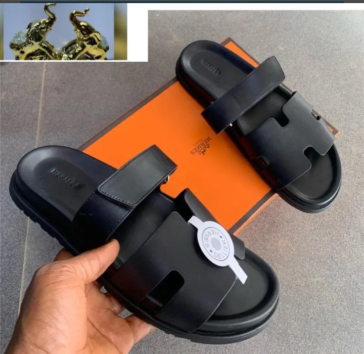 A pair of luxurious black leather slide sandals with double strap and silver accent, held by a hand against an orange shoe box, exuding premium quality and style.