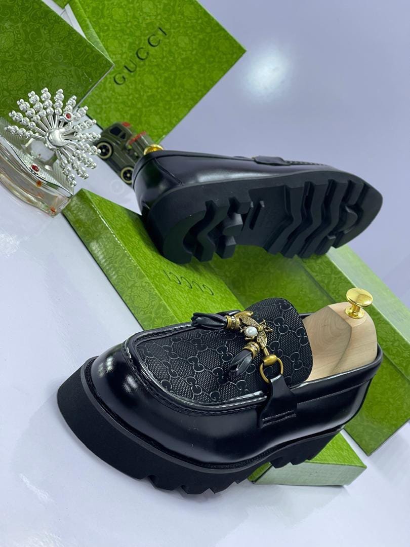 A pair of black loafers with gold accents and Gucci-inspired design, placed on a white surface with green shoe boxes in the background.