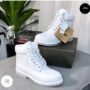 A pair of premium white leather combat boots with lug sole, displayed on a table with a shoe box and other luxury items in the background.