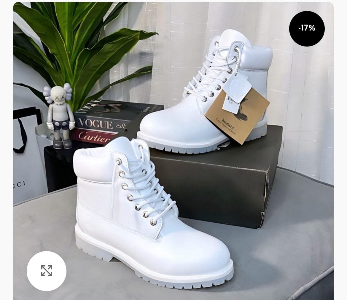 A pair of premium white leather combat boots with lug sole, displayed on a table with a shoe box and other luxury items in the background.