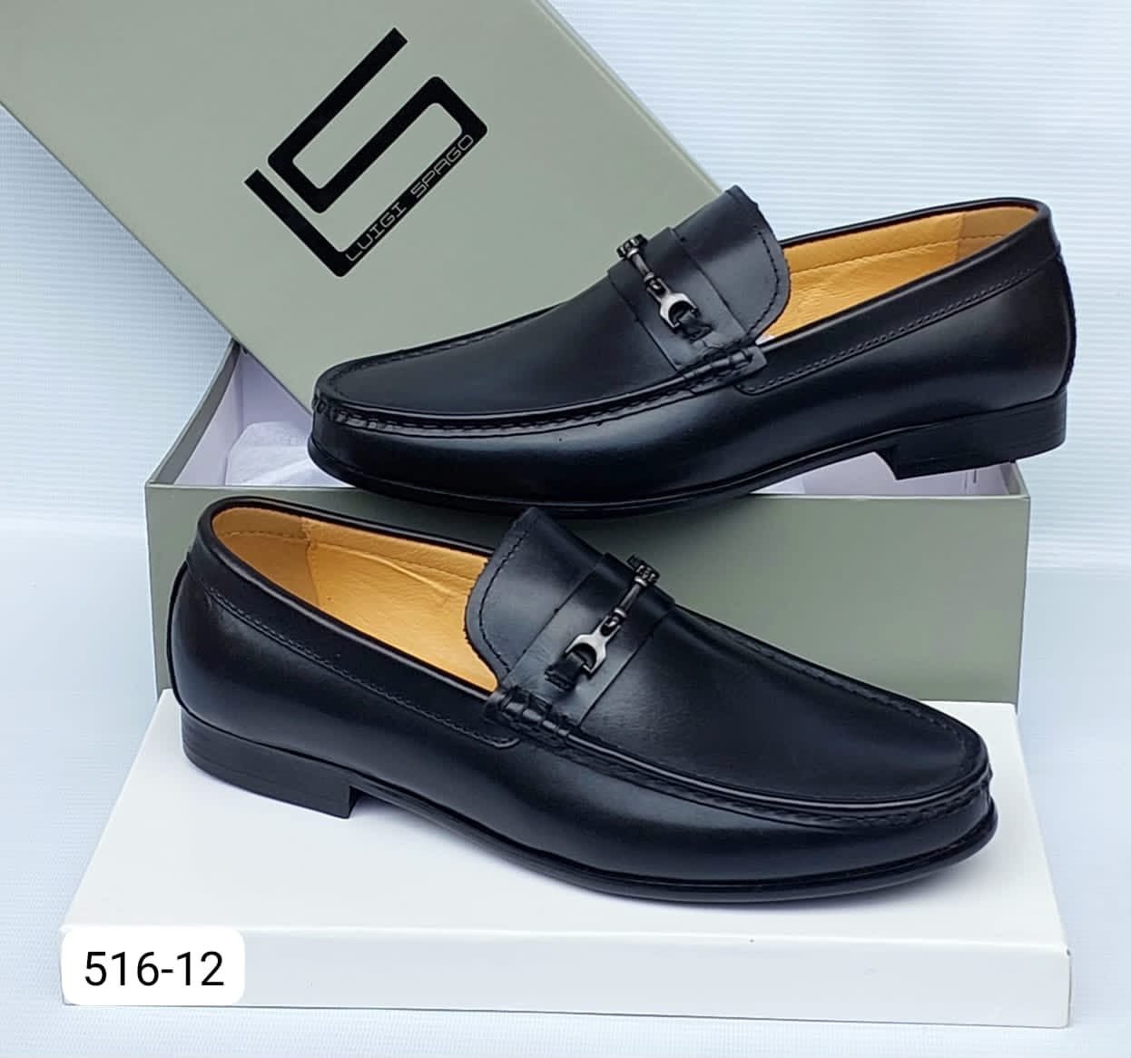 A pair of premium black leather loafers with sleek buckle detail, displayed on a white box with a grey shoe box in the background.