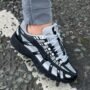 A close-up shot of a person wearing black and white Nike F-6000 series sneakers on a rocky surface, highlighting the shoe's sleek design and comfort.