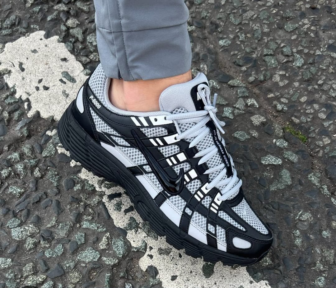 A close-up shot of a person wearing black and white Nike F-6000 series sneakers on a rocky surface, highlighting the shoe's sleek design and comfort.