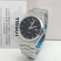 A sleek Tissot PRX watch with a black face and silver metal band, presented with a 2-year international warranty card.