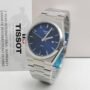 A sleek Tissot PRX watch with a blue face and silver band, wrapped in plastic and displayed in front of a Tissot warranty card.