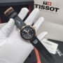 A luxurious Tissot watch with a black leather strap and gold accents, displayed on a white-gloved hand with its packaging in the background.