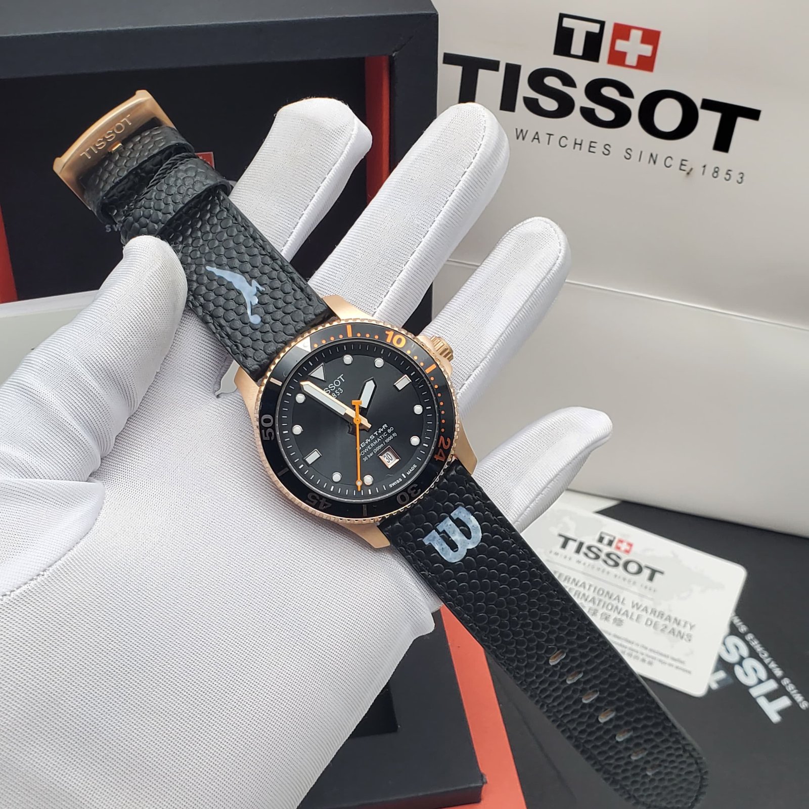 A luxurious Tissot watch with a black leather strap and gold accents, displayed on a white-gloved hand with its packaging in the background.