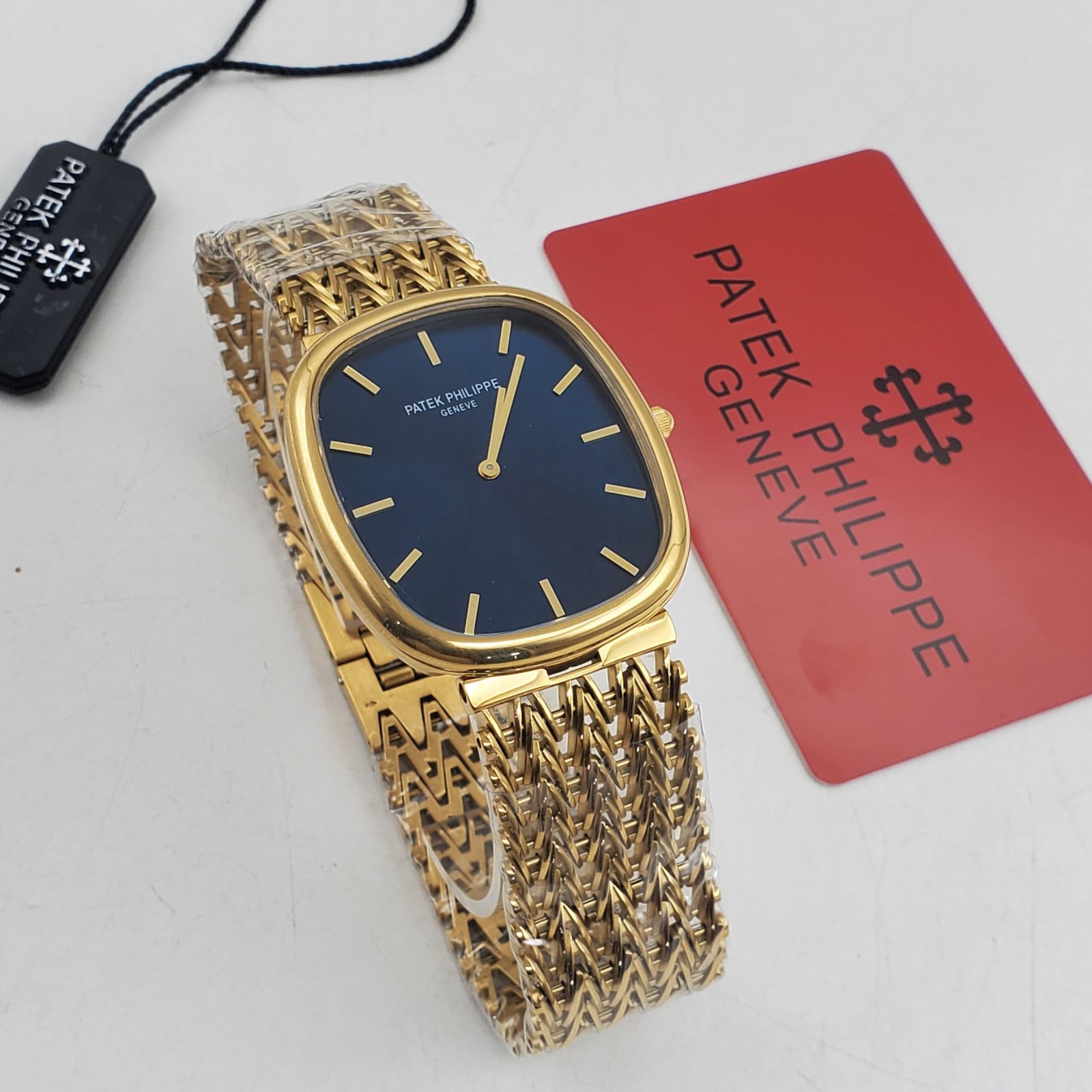 A luxurious gold Patek Philippe watch with a blue face and gold band, displayed on a white surface with a red card and black tag.