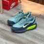 A pair of Nike sneakers in light blue with dark blue and neon green accents, positioned on a wooden floor next to a red Nike shoe box.