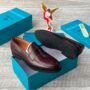 A pair of luxurious brown leather loafers displayed on a teal shoe box, with the box and a decorative angel figurine in the background.